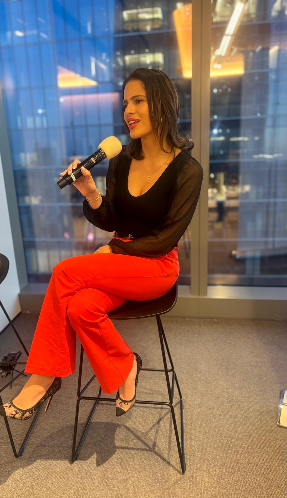 Jazzlyn Feliciano speaking on a stool with the NYC skyline behind her